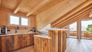 A modern kitchen with wooden beams, a large island, and a sliding glass door leading to an outdoor area. - Chalet Sunshine – Alpine Charm With Stunning Views (Garmisch-Partenkirchen)