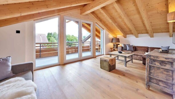 A spacious loft with wooden beams, a large sliding glass door leading to a balcony, a cozy seating area with a sofa and armchairs, and a rustic wooden table. - Chalet Sunshine – Alpine Charm With Stunning Views (Garmisch-Partenkirchen)
