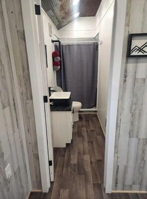 Un couloir étroit, doté d’un plancher en bois, mène à une salle de bain. La salle de bain est équipée de toilettes, d’un lavabo et d’une douche avec un rideau.
