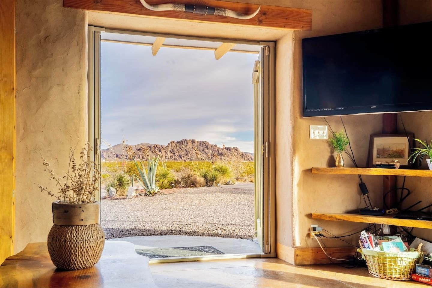 Une chambre avec une porte donnant sur un paysage désertique, une télévision et une étagère contenant des livres et des plantes.