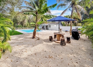 A pool area with a circular table and chairs, surrounded by sand and palm trees.