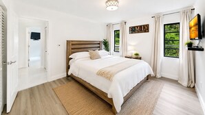A bedroom with a large bed, a bedside table with a lamp, a TV mounted on the wall, and a window with curtains. - Charming Nature Farmhouse Getaway – Spacious, Peaceful, Relaxing (Miami)