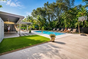 A backyard with a swimming pool, a hammock, lounge chairs, and a basketball hoop.