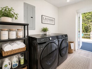 A laundry room with two black Samsung washing machines, shelves with detergent and fabric softener, and a sign that says 'LAUNDRY WASH DRY FOLD REPEAT'.