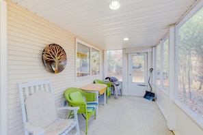 Relax on the covered porch with comfortable wicker chairs and a wooden table, surrounded by a serene wooded view.