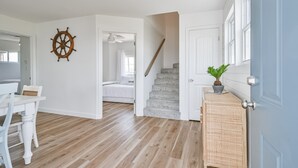 A room with a wooden floor, a bed, a white desk, a chair, a wall-mounted ship's wheel, a staircase, and a door with a potted plant. - NEW! Bayfront home w/ private pier, steps to beach (Freeport)