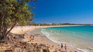 A beach with clear turquoise waters, rocky shoreline, and a few people walking along the shore. - Barefoot Buddina Laid-Back Pet Friendly by the Sea (Buddina)