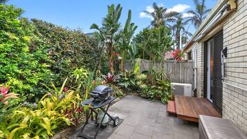 A patio with a barbecue grill, a wooden bench, and various tropical plants.