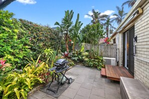 A patio with a barbecue grill, a wooden bench, and various tropical plants.