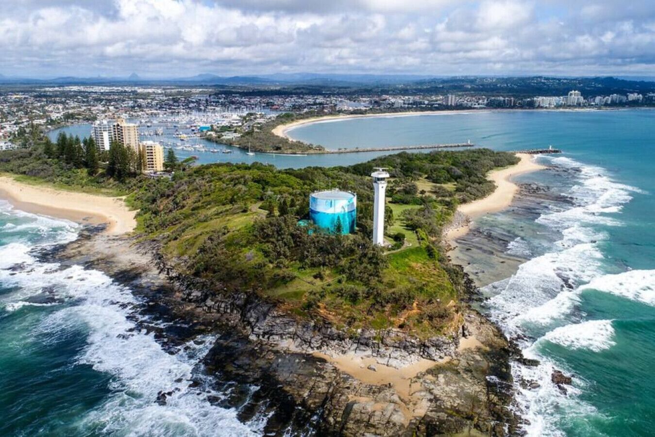 Une vue aérienne d’une zone côtière, avec un phare, une plage et une vue sur la ville en arrière-plan.