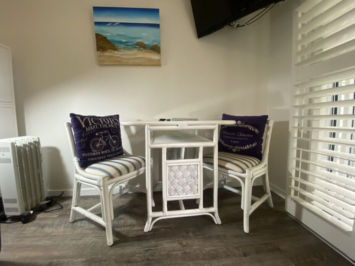 Une pièce d’angle avec une chaise et une table en osier peintes en blanc, un radiateur et une fenêtre avec des stores.