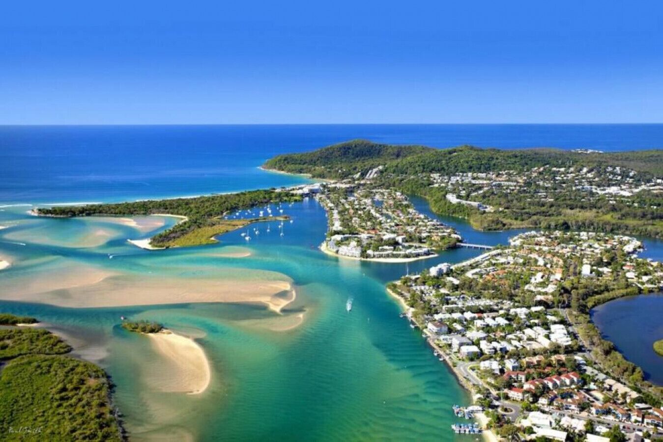 An aerial view of a coastal town with clear blue waters, sandy beaches, and a dense residential area with houses and buildings.
