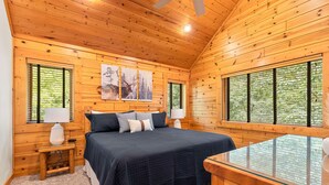 A wooden cabin bedroom with a bed, two bedside tables, a glass table, and large windows with blinds.