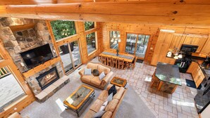A log cabin interior with a stone fireplace, wooden furniture, and large windows.