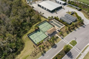 Vue aérienne d’un complexe comprenant un court de tennis, un espace de repos couvert et des bâtiments adjacents.