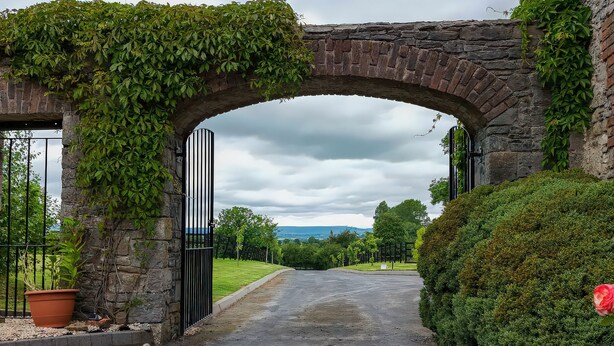 A stone archway leading to a paved path, surrounded by greenery and a potted plant.