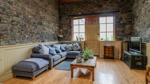 A cozy living room with a stone wall, a wooden ceiling, a grey sectional sofa, a wooden coffee table, and a television on a cabinet.