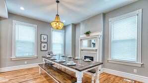 A dining room with a wooden table set for six, flanked by white-framed windows, a fireplace, and a hanging light fixture.
