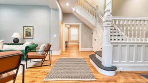 A spacious living room with a staircase leading to an upper floor, a green sofa, a patterned rug, and a framed picture on the wall.