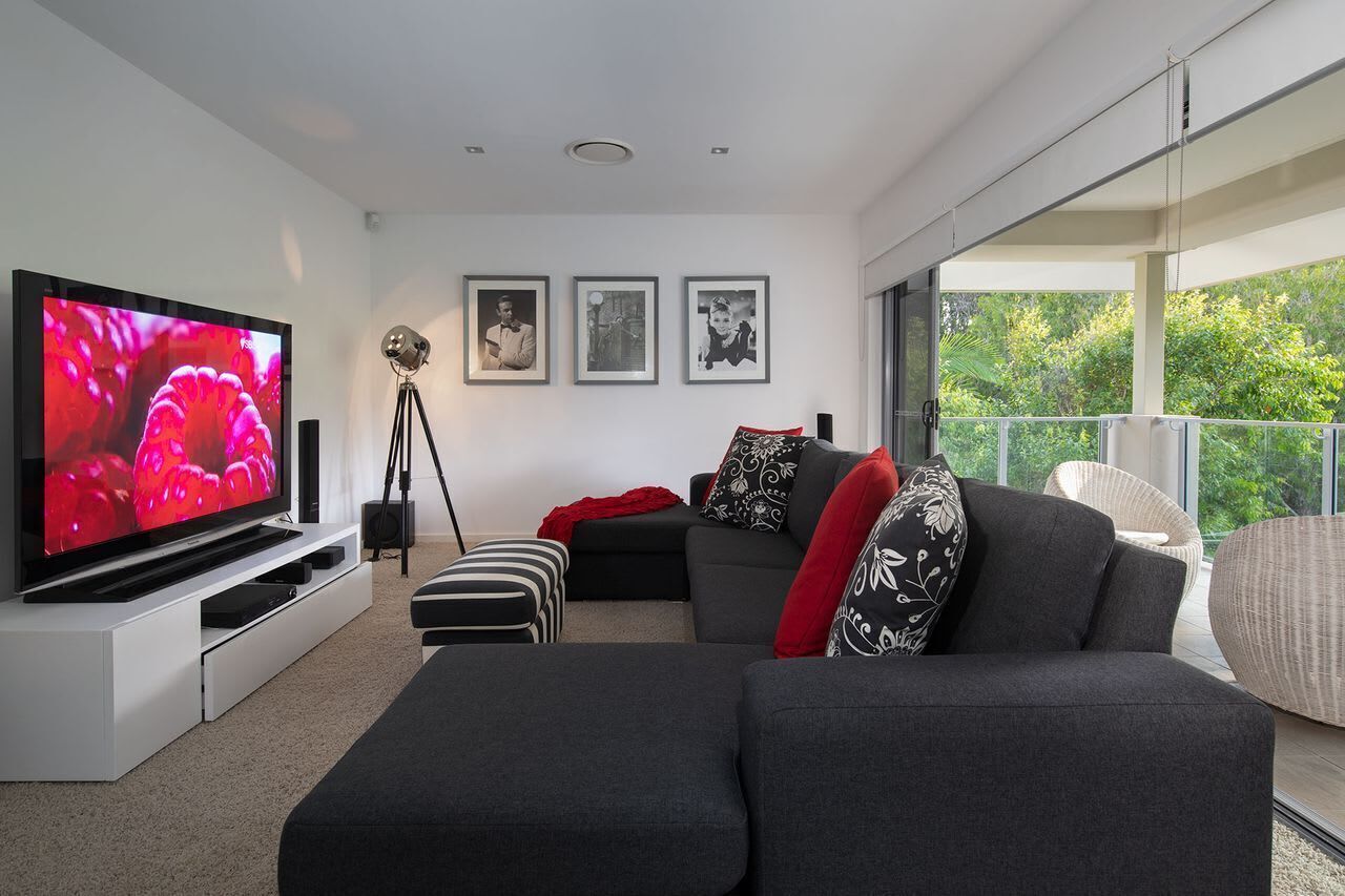 A modern living room with a large flat-screen TV, a sofa, a chair, and a balcony with a view of greenery.