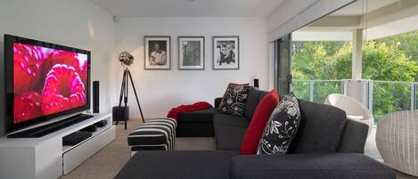 A modern living room with a large flat-screen TV, a sofa, a chair, and a balcony with a view of greenery.