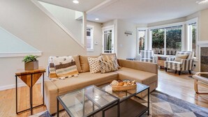 A modern living room with a sofa, coffee table, and striped armchairs. A staircase leads to another level, and there is a large window with a view of greenery outside. - Perch - Walk to Lake Union! Spacious 3BR/3.5BA w/ Garage (Seattle)