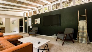 Una sala de estar moderna con un sofá modular, una mesa de centro y dos sillones. Un televisor de pantalla plana grande está montado en la pared, y una escalera está apoyada contra la pared.