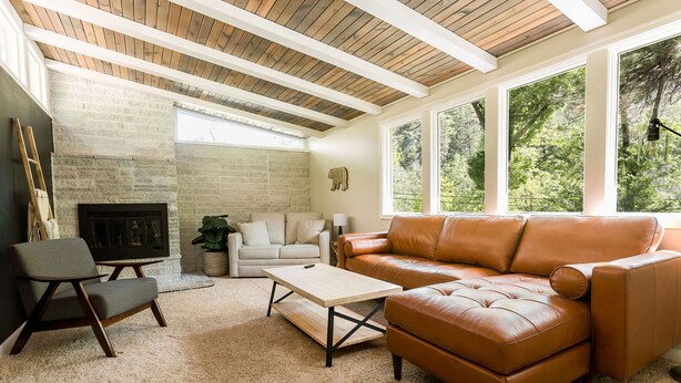 Una sala de estar con chimenea de piedra, un sofá de cuero, una mesa de centro de madera y ventanas grandes que ofrecen vistas a la vegetación.