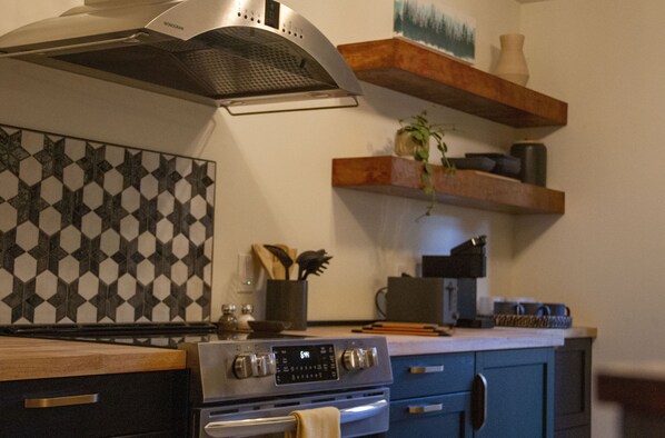 Une cuisine équipée d’une cuisinière, d’un four et d’un crédence ornée d’un motif géométrique. Des étagères en bois, situées au-dessus du plan de travail, servent à ranger divers ustensiles de cuisine.