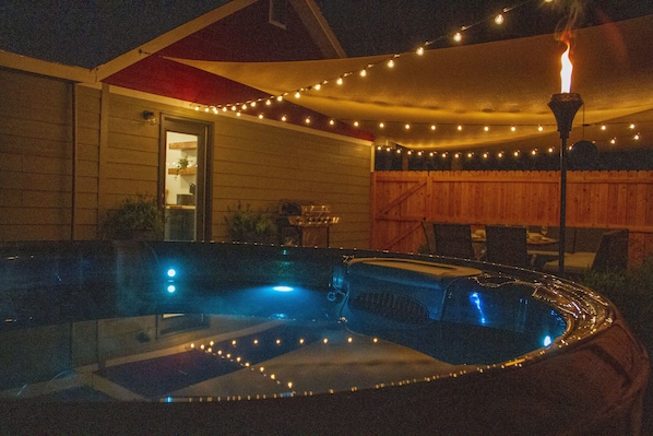 A hot tub with string lights and a fire feature, surrounded by a wooden fence and a covered patio area. - Brand New - Gracious Old Town Guest House w/ Hot Tub (Fort Collins)