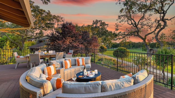 Una terraza con mobiliario de mimbre, una mesa redonda con bebidas y vistas a un parque y al atardecer.