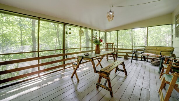 Una terraza de madera con bancos y mesas de picnic, ventanas amplias con vistas al bosque y una lámpara colgante.