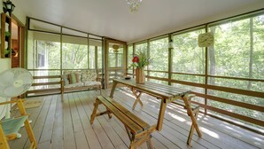 Una terraza de madera con bancos y un ventilador, amplias ventanas con vistas a árboles, y una lámpara colgante.