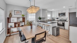 A modern kitchen with white cabinets, a wooden dining table, and a hanging light fixture.