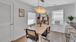 A modern dining area with a wooden table, black and wicker chairs, a hanging light fixture, and a kitchen counter with stools.