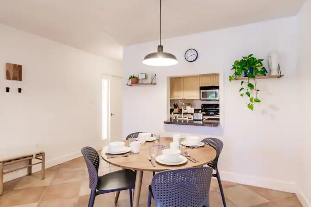 A dining area with a round table set for four, surrounded by chairs. A kitchen is visible in the background with wooden cabinets and a microwave.