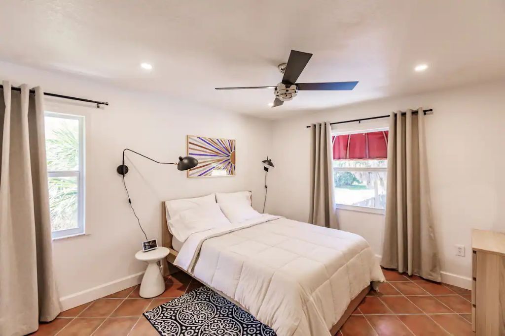 A bedroom with a bed, a ceiling fan, a nightstand, a lamp, a window with curtains, and a wall art piece.