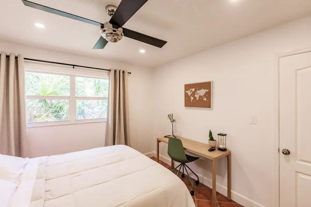 A bedroom with a bed, a desk, a chair, a ceiling fan, a window with curtains, and a door.