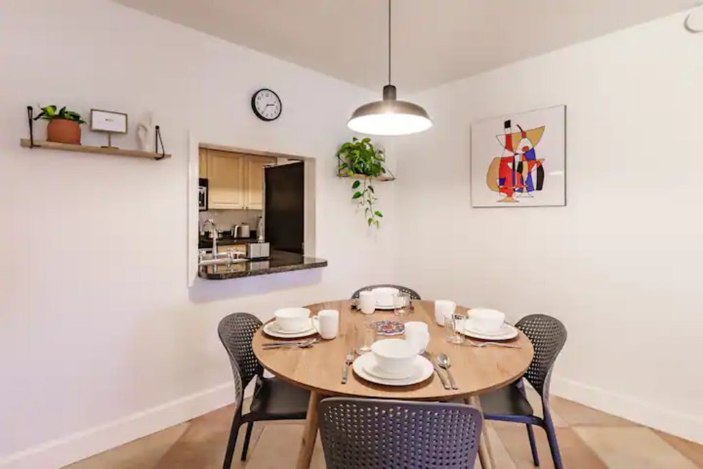 Un comedor con una mesa redonda dispuesta para cuatro personas, rodeada de sillas negras. Hay una planta colgante, un reloj y una obra de arte enmarcada en la pared.