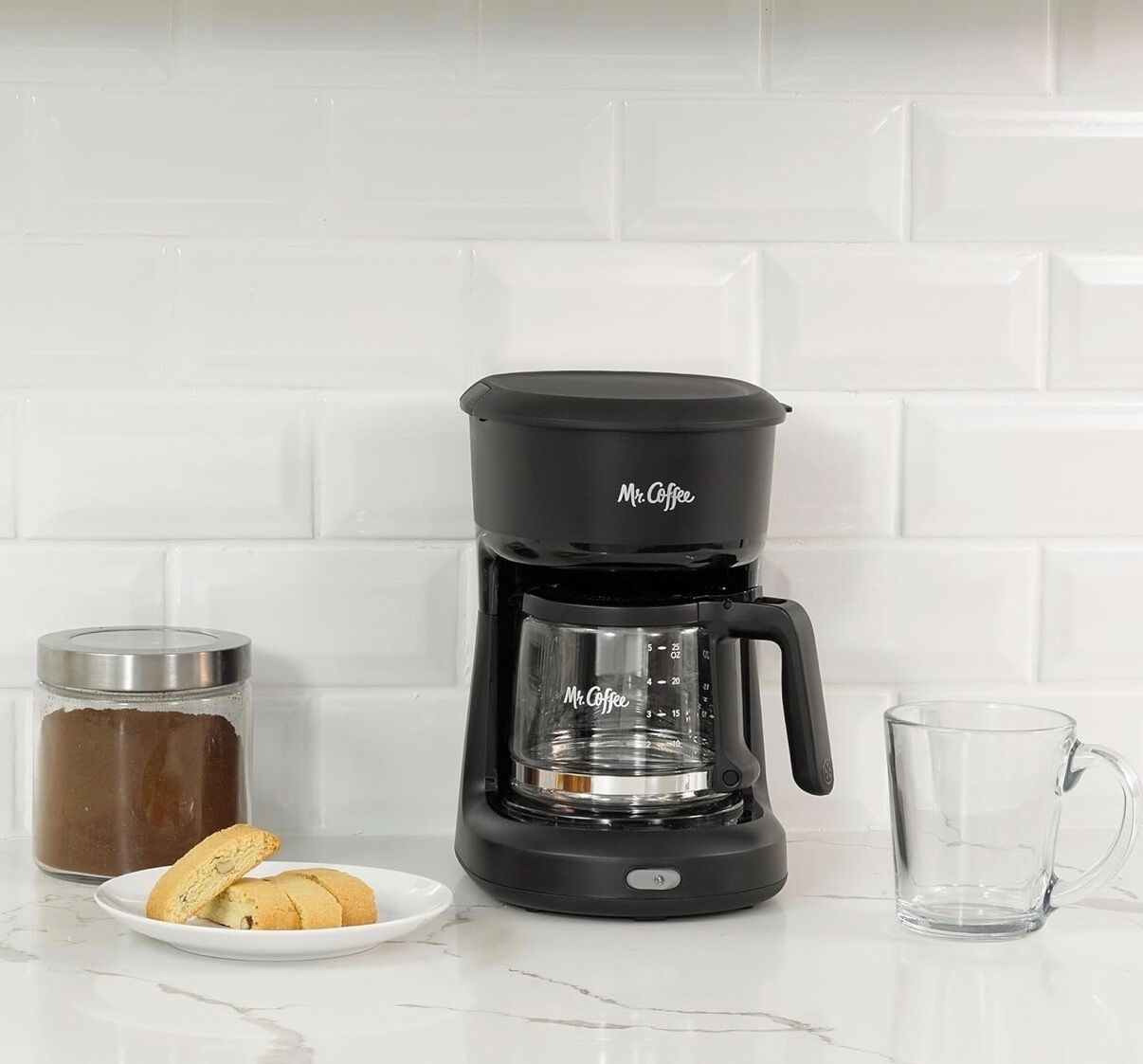 Une cafetière noire de la marque Mr. Coffee, posée sur un comptoir blanc, avec une tasse en verre et un bocal de café.