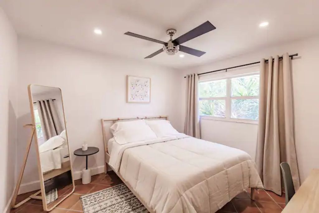 A bedroom with a bed, a ceiling fan, a window with curtains, a bedside table, and a mirror.