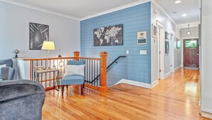 Una sala de estar con una pared azul de acento, piso de madera, una escalera con barandal de madera y un escritorio con lámpara.