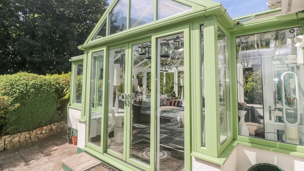 A conservatory with a green frame, large glass doors, and a view of the garden. - Cornerstone (Penzance)