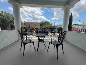 A covered balcony with a table and chairs, white columns, and a view of buildings and trees.