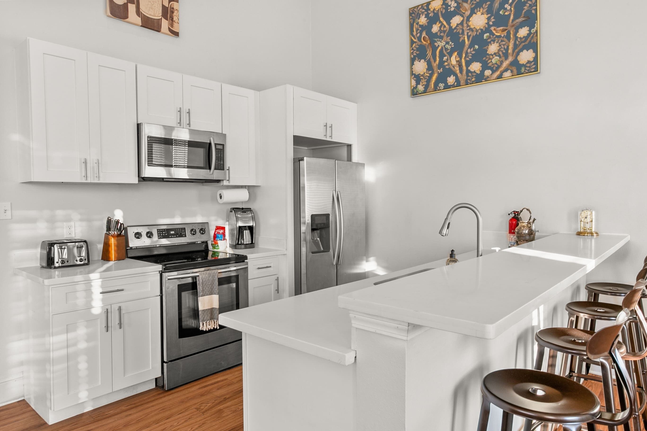 A modern kitchen with white cabinetry, stainless steel appliances, a central island with stools, and a decorative wall piece.
