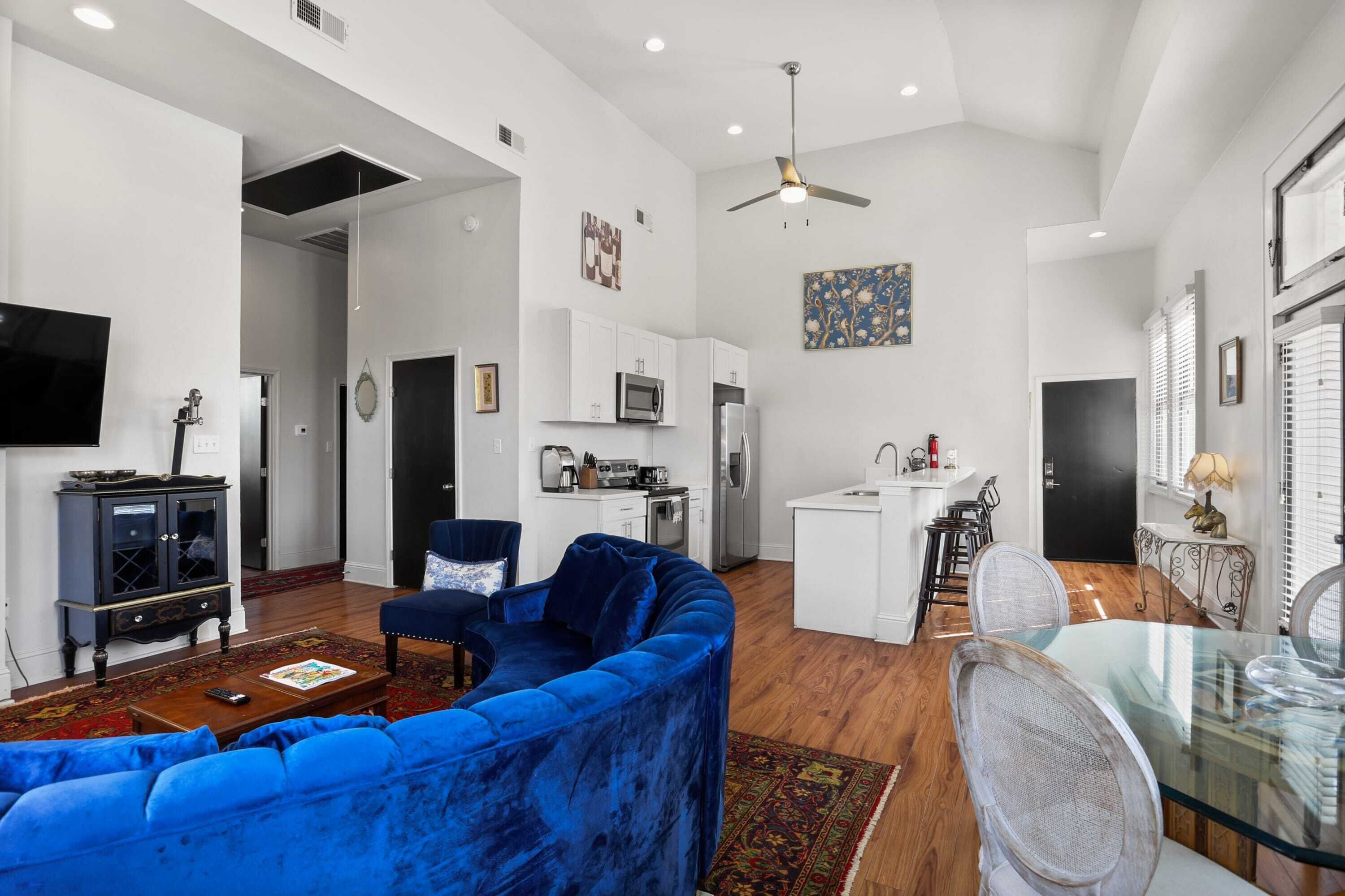 A modern living room with a blue sofa, a glass dining table, and a kitchen in the background.