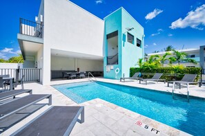 Ein Schwimmbecken mit Liegestühlen und einem Tisch, umgeben von einem Gebäude mit einem Balkon und einem Schild, das eine Wassertiefe von 1,5 Metern angibt.