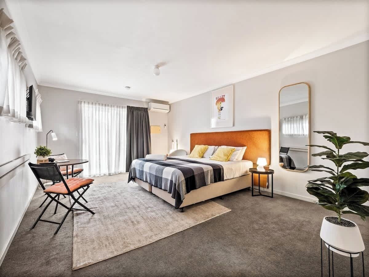 The photo showcases a spacious, modern studio with a plush bed, stylish orange headboard, bedside tables, a large mirror, cozy seating area, air conditioning, natural light, and sliding doors to a private space.