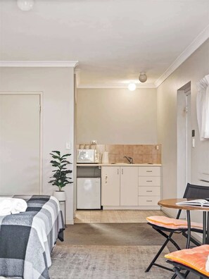 A compact room with a bed, a small kitchenette, a dining table with two chairs, and a potted plant.