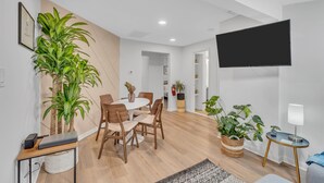 A modern living room with a dining area, a flat-screen TV mounted on the wall, and several potted plants.